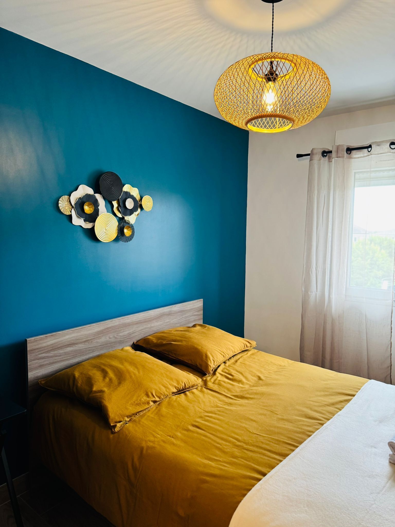 Une chambre avec un mur bleu, une tête de lit en bois, un lit avec une literie jaune, un luminaire suspendu et une fenêtre avec des rideaux.
