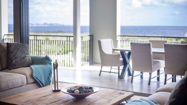 Una sala de estar moderna con una mesa de centro de madera, un bol de aperitivos y vistas al océano a través de amplios ventanales.