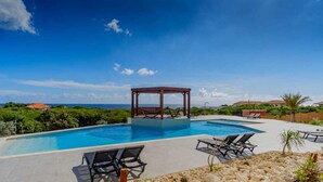Una piscina con sillas de descanso, un pabellón y vista al océano.