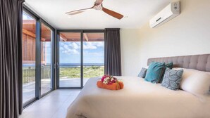 Habitación de hotel con una cama grande, ventilador de techo, aire acondicionado y vistas al océano a través de amplias ventanas.