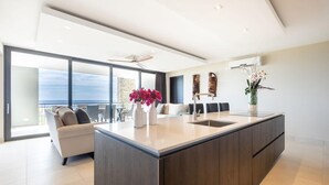 Una cocina moderna con una isla grande, un área de comedor con sofá y sillas, y vista al océano a través de puertas correderas amplias.