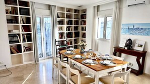 Een eetkamer met een houten tafel voor vier personen, omringd door witte stoelen. Links bevindt zich een boekenplank vol boeken, en rechts een schilderij aan de muur.
