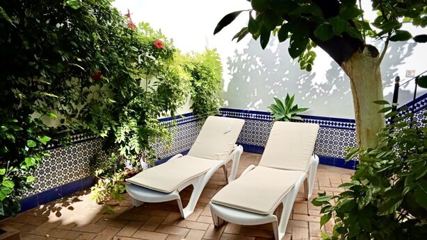 Een terras met twee witte lounge fauteuils, een betegelde vloer en een muur versierd met blauw-witte tegels en groene planten.