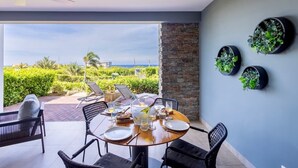 A dining area with a round table set for four, surrounded by wicker chairs, with a view of a lush garden and the sea. - Luxury ground-floor retreat with poolside terrace - Coral Estate (Sint Willibrordus)