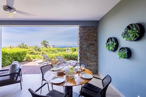 Ein Essbereich mit einem runden Tisch, gedeckt für vier Personen, umgeben von Korbstühlen, mit Blick auf einen üppigen Garten und das Meer.