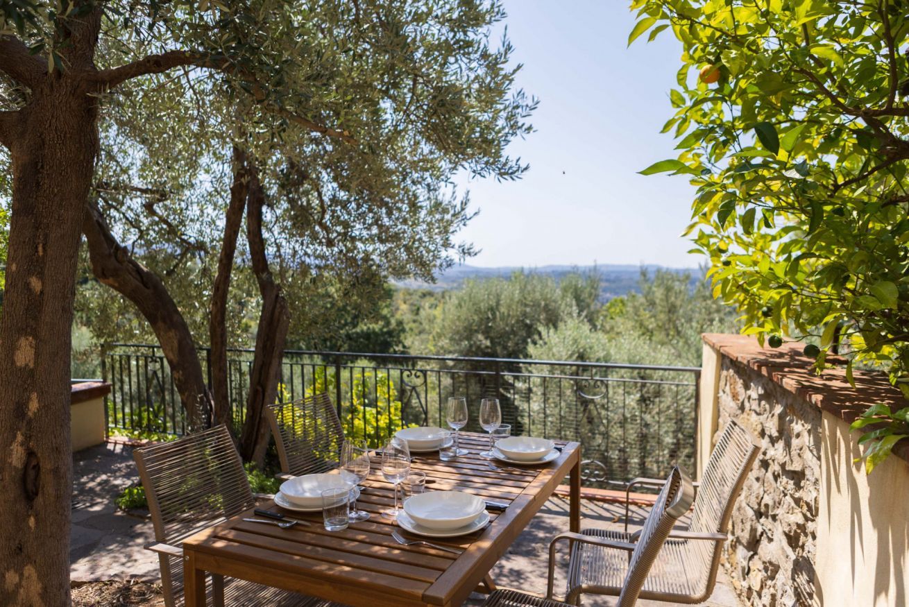 An outdoor dining area with a wooden table set for four, surrounded by wicker chairs, overlooking a scenic landscape.