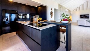A modern kitchen with dark cabinetry, a central island, and a dining area with a white sofa and a TV. - Luxury penthouse with panoramic sea views - Coral Estate (Sint Willibrordus)