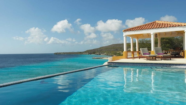 Una piscina infinita con agua azul cristalina, un pabellón blanco con detalles amarillos y vistas al océano y la tierra distante.
