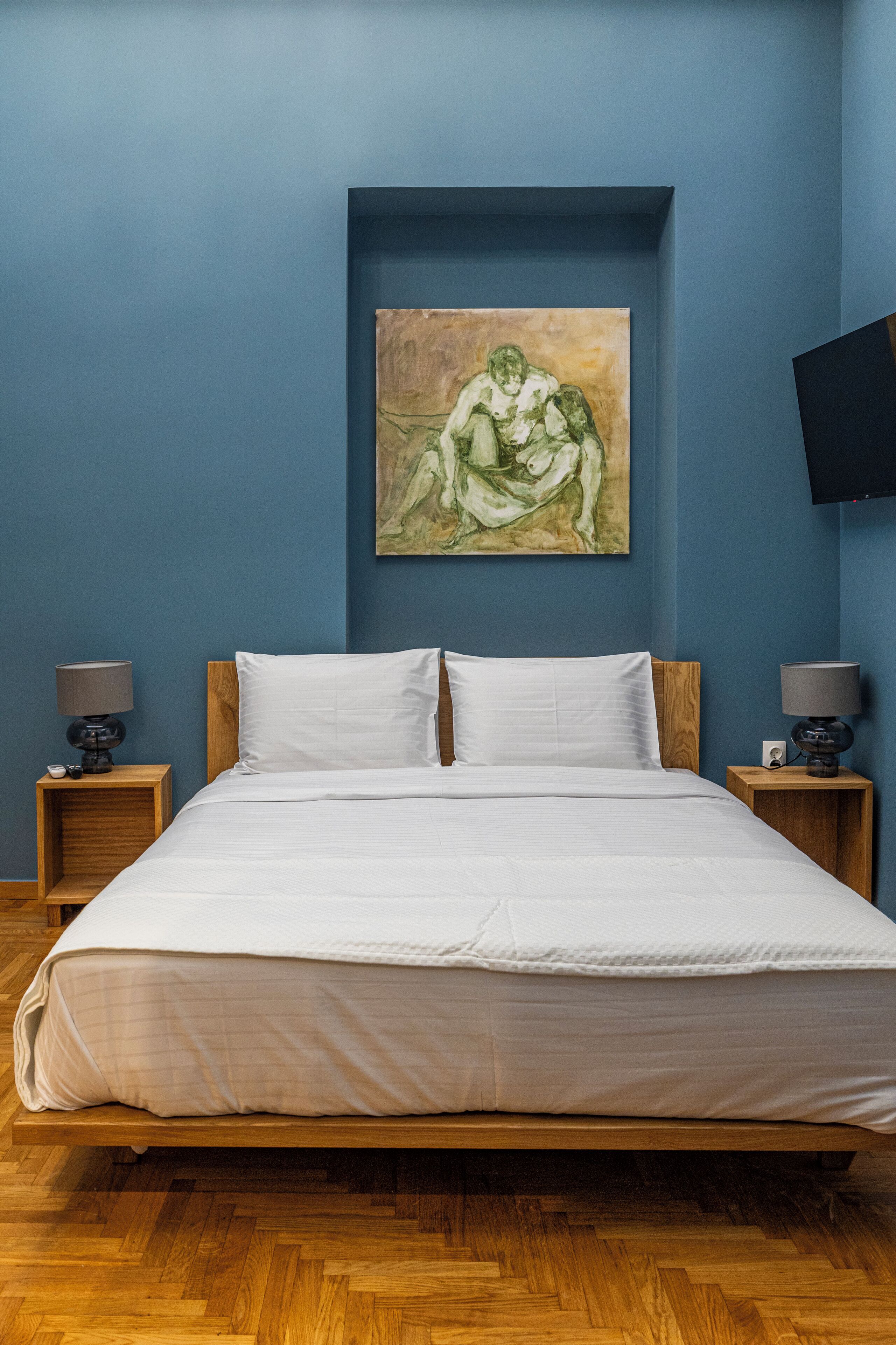 A bedroom with a bed, two nightstands, a painting on the wall, and a flat-screen TV mounted above the bed.