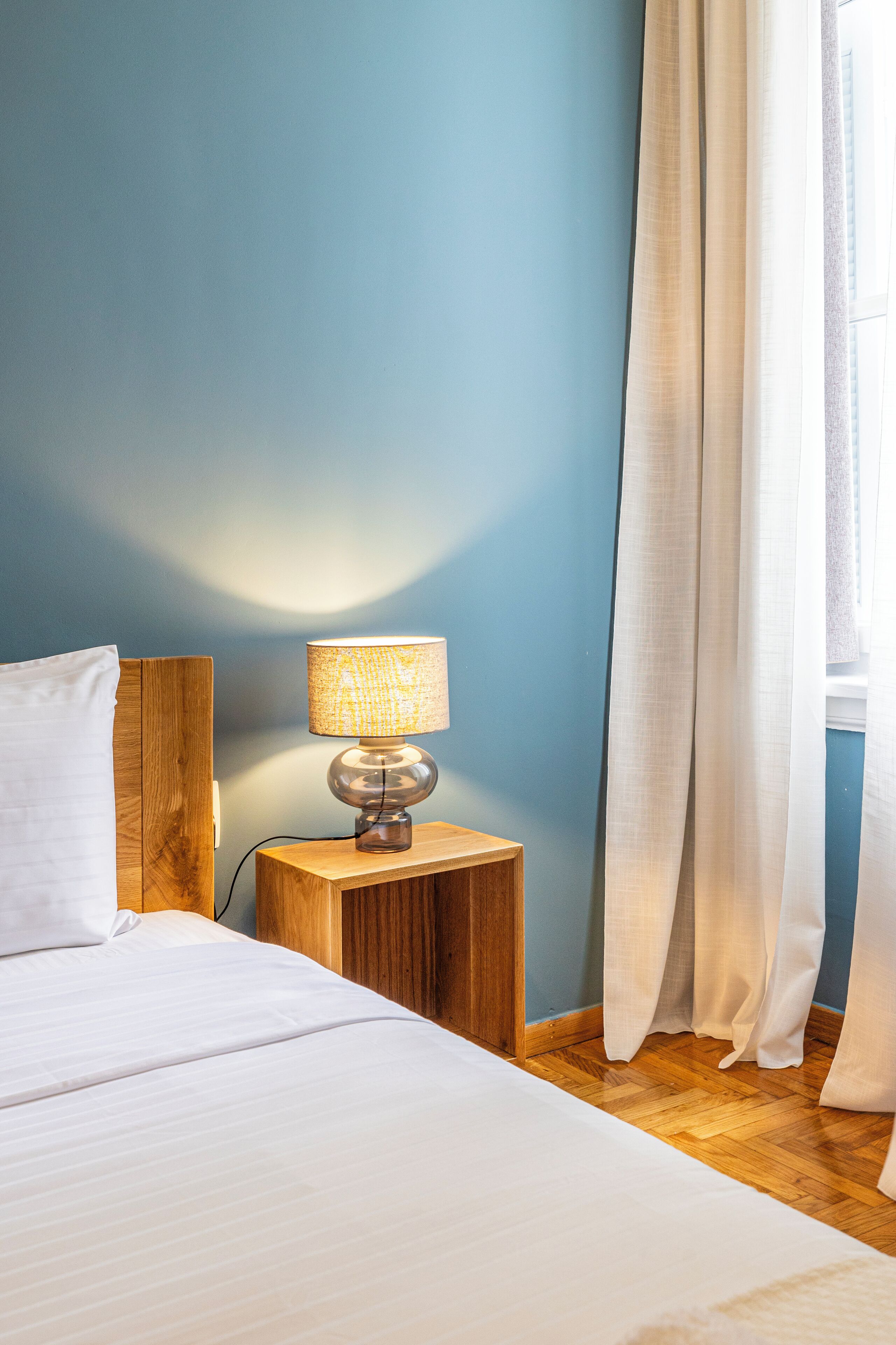A neatly made bed with white linens, a wooden nightstand with a lamp, and a window with white curtains.