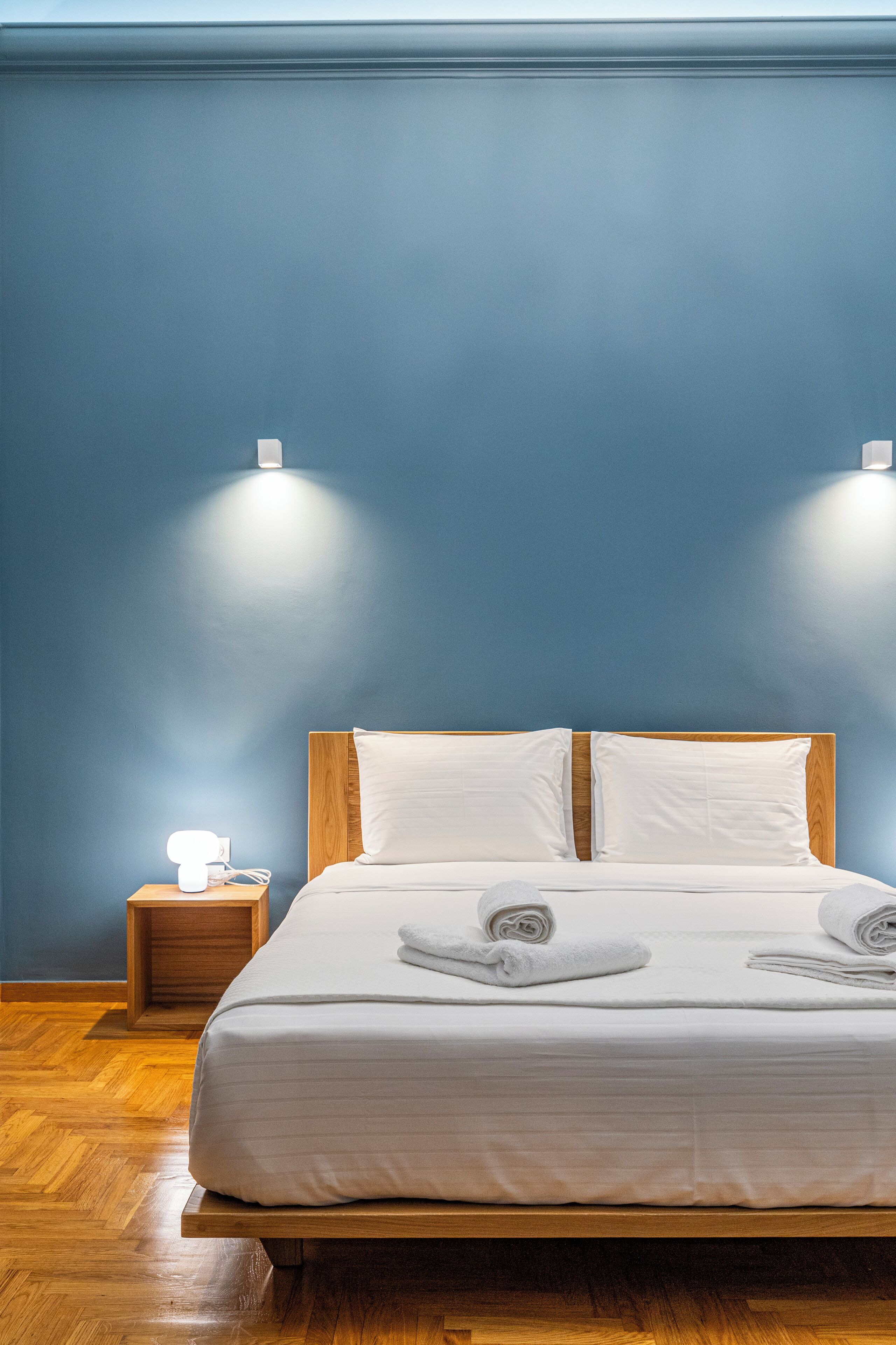 Una cama bien hecha con sábanas blancas y dos almohadas, una mesita de noche de madera con una lámpara, y una pared azul.
