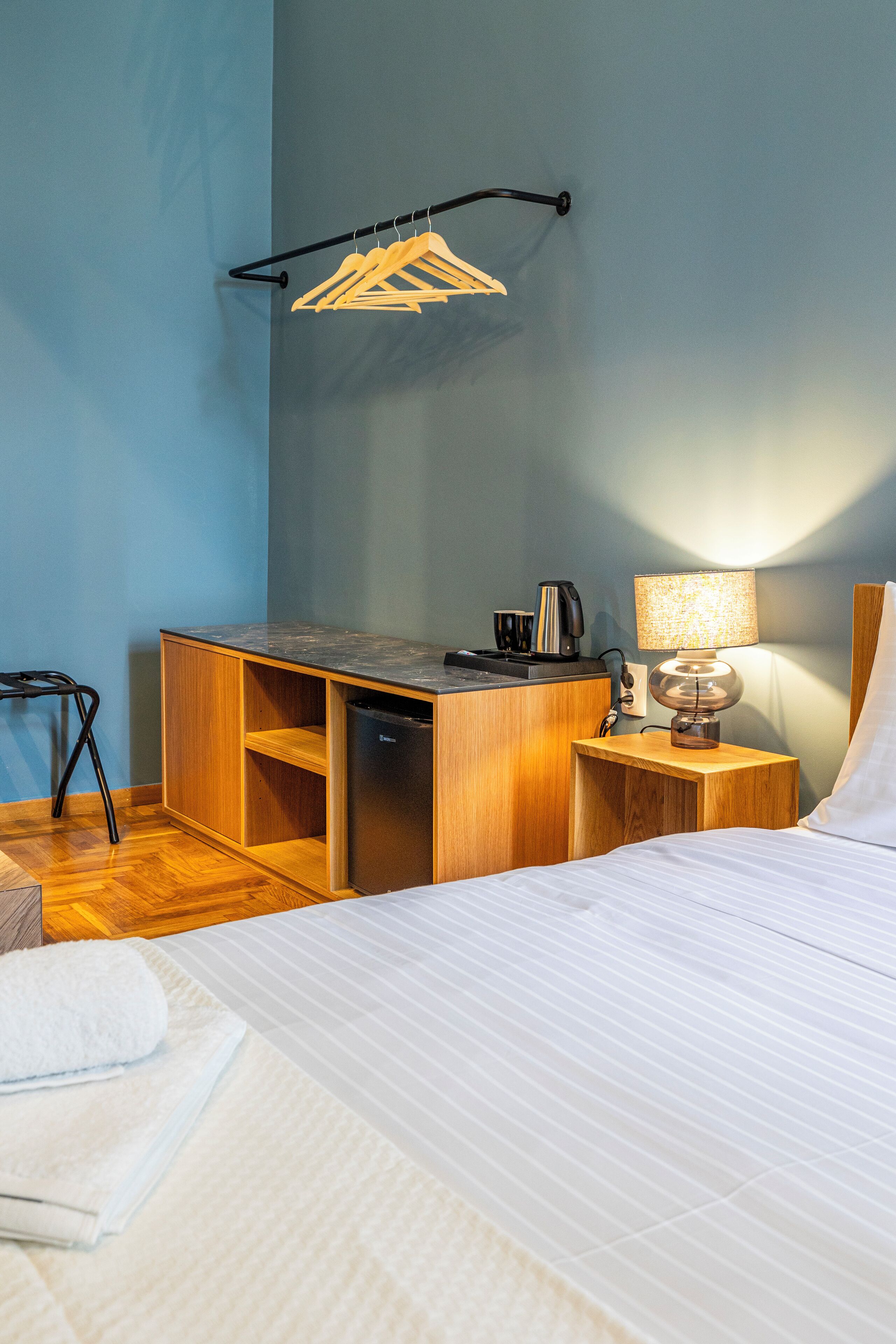 A compact hotel room with a bed, a desk with a computer, a chair, a small table with a lamp, and a coat rack with hangers.