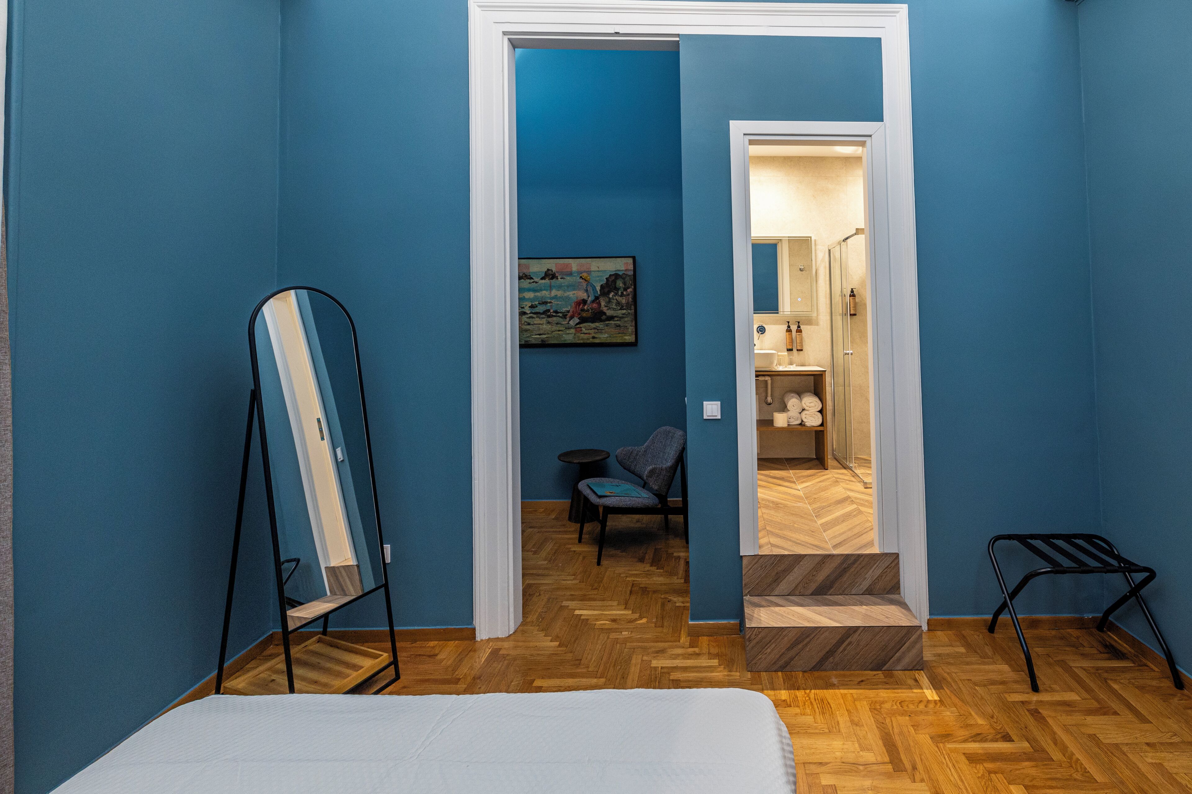 Una habitación con paredes azules, suelo de madera en espiga, una cama, una silla, una mesita y un cuadro en la pared.