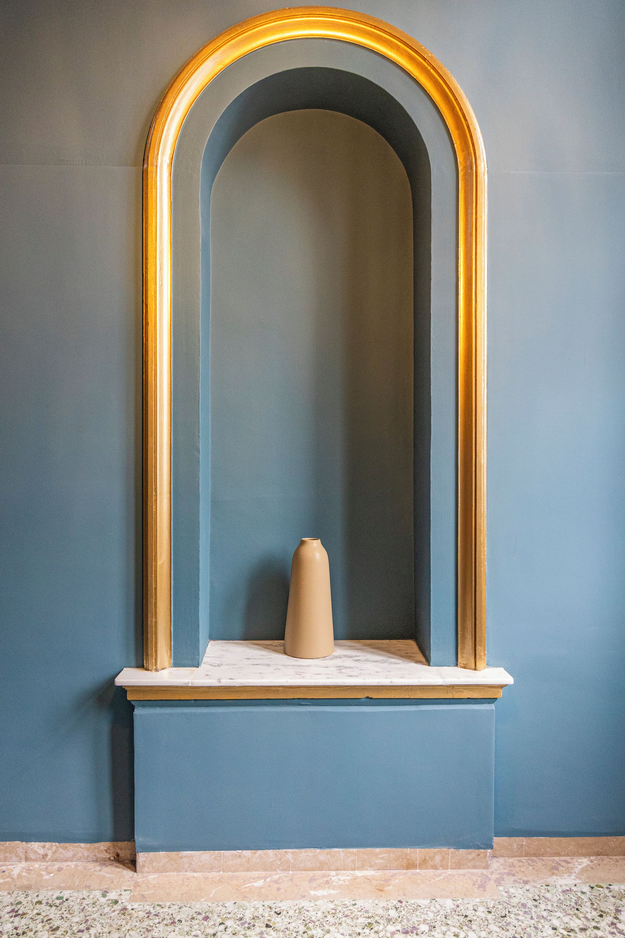 A room with a blue wall, a marble floor, and a beige vase on a marble pedestal.