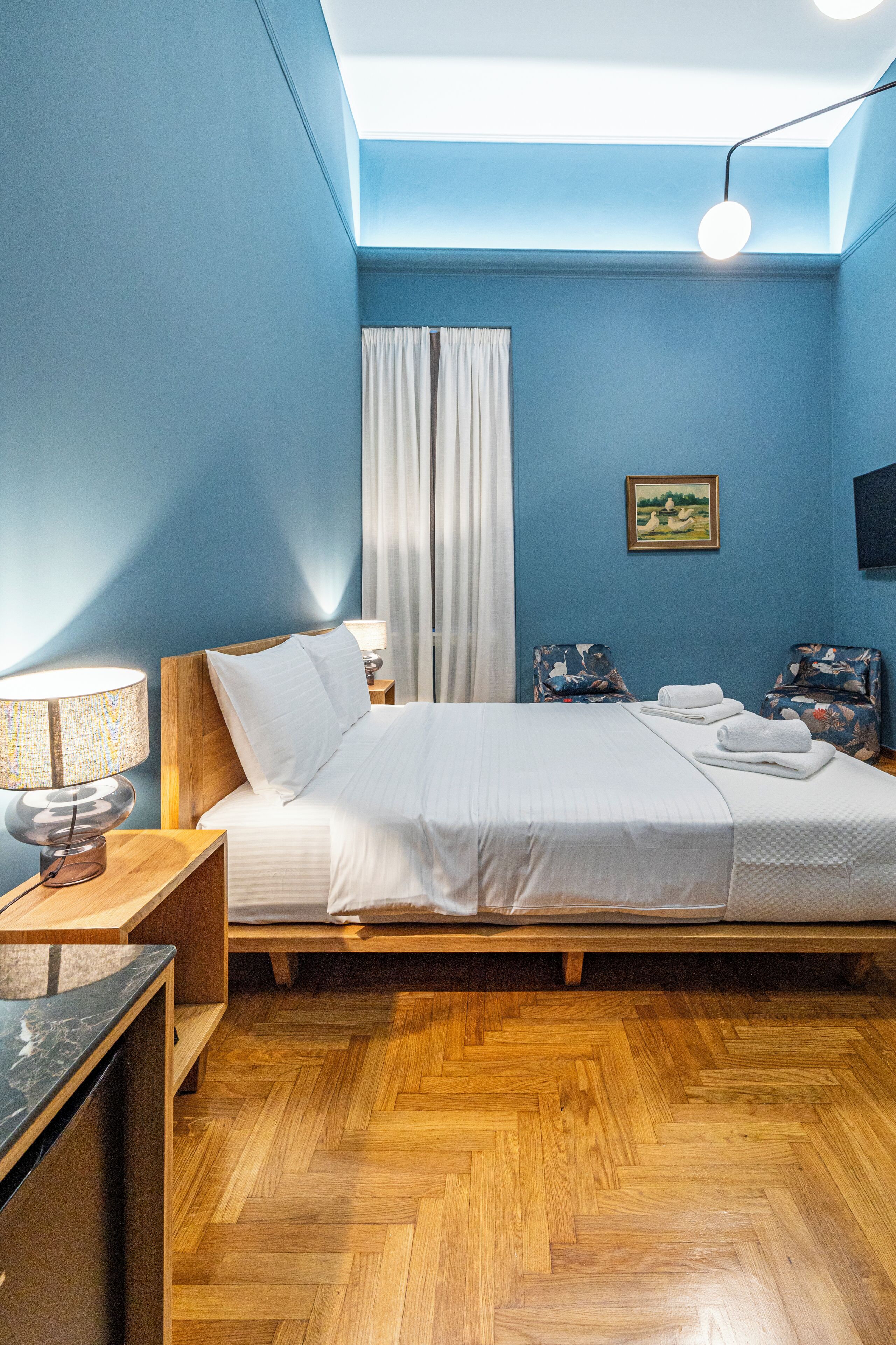 Un dormitorio con cama de madera, una mesita de noche con lámpara, una silla con cojín floral y un cuadro en la pared.