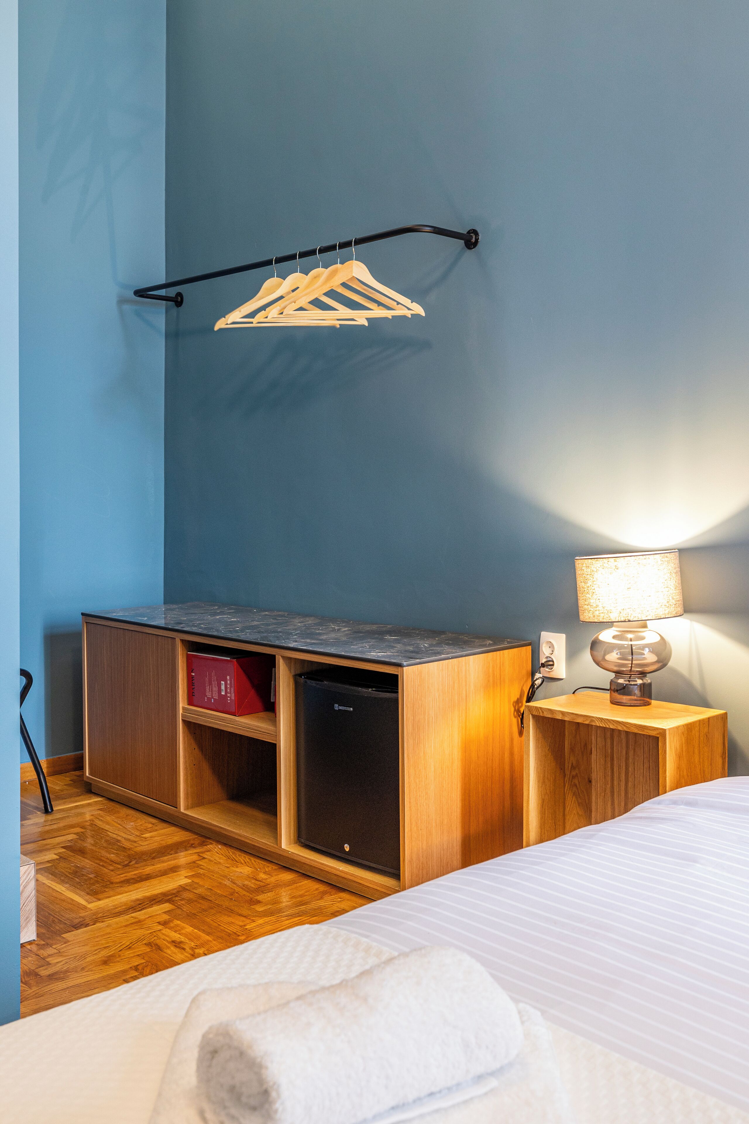 A hotel room with a bed, a wooden dresser, a desk with a lamp, and a clothes rack.