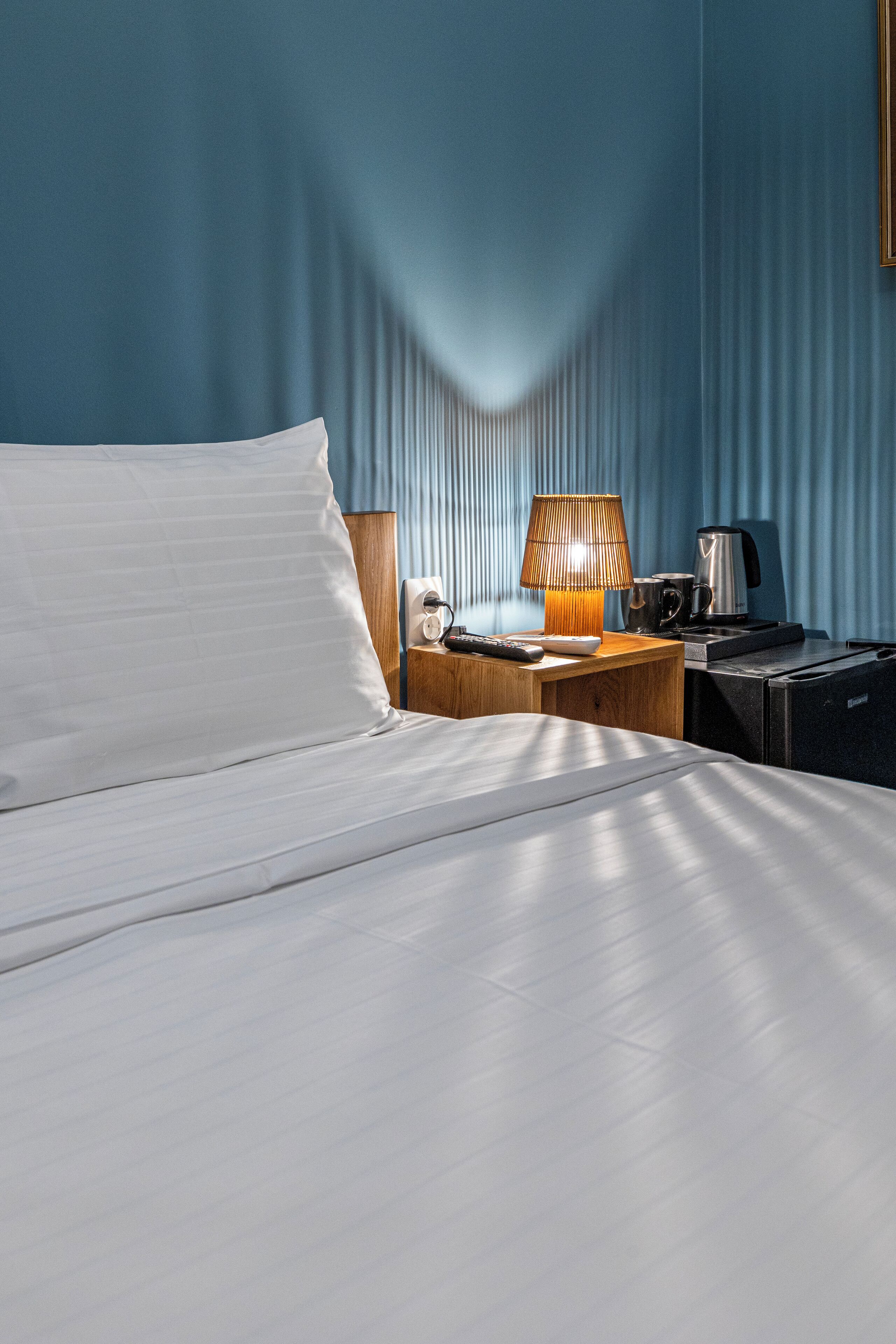 A hotel room with a neatly made bed, a wooden nightstand with a lamp, a coffee maker, and a small table with a remote control.