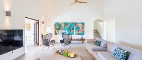 Amplia sala de estar con techo alto, ventilador de techo, un cuadro abstracto de gran tamaño, un conjunto de sofás y una mesa de centro con objetos decorativos.
