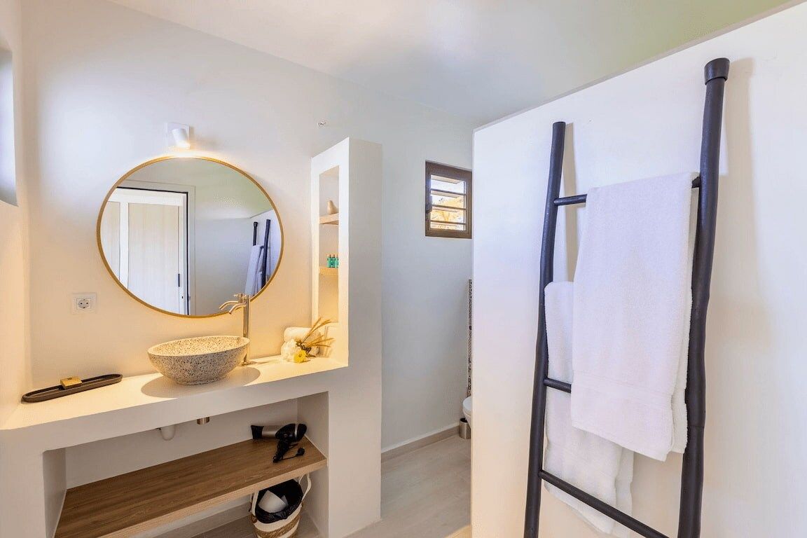 A bathroom with a round mirror, a bowl sink, a towel rack, and a shelf with toiletries.