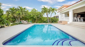 Une piscine rectangulaire aux carreaux bleus, entourée de palmiers et bordée d’un bâtiment blanc doté d’un toit rouge.