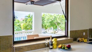 Un comptoir de cuisine donnant sur une piscine et des arbres, une bouteille de champagne et un saladier rempli de fruits.
