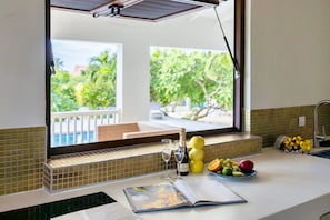 A kitchen counter with a view of a pool and trees, a bottle of champagne, and a bowl of fruit. - Stylish retreat with tropical garden and pool (Sint Willibrordus)