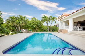 Una piscina con borde de azulejos, rodeada de palmeras y un edificio blanco con balcón.