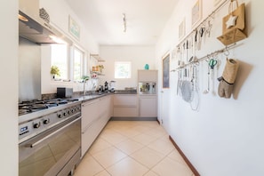 Une cuisine compacte équipée d’une cuisinière à gaz, d’un four et d’un réfrigérateur. Des ustensiles de cuisine sont accrochés au mur, et une fenêtre apporte de la lumière naturelle.