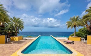 A rectangular pool with a clear blue center, surrounded by yellow walls and palm trees, leading to the ocean. - Spacious villa with infinity pool and ocean view (Sint Willibrordus)