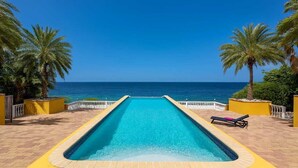Uma piscina retangular com superfície de água azul cristalina, rodeada por paredes amarelas e palmeiras, com vista para o oceano.