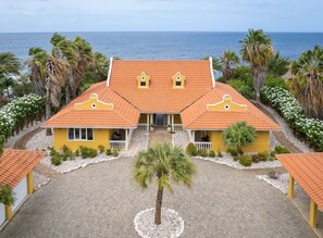 A two-story building with a symmetrical facade, a central entrance, and a paved courtyard. - Spacious villa with infinity pool and ocean view (Sint Willibrordus)