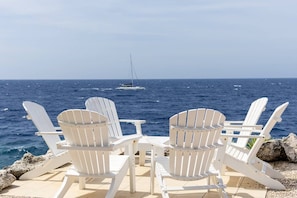 Deux chaises Adirondack blanches sont placées face à une étendue d’eau, avec un voilier visible au loin.