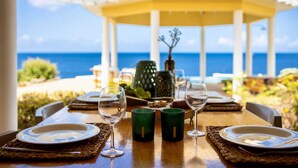 Une table à manger dressée pour deux personnes, avec des assiettes, des verres et une bougie verte, donnant sur l’océan.