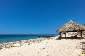 A beach with a thatched-roof hut, a bench, and two lounge chairs. - Oceanfront villa - infinity pool and private beach (Sint Willibrordus)