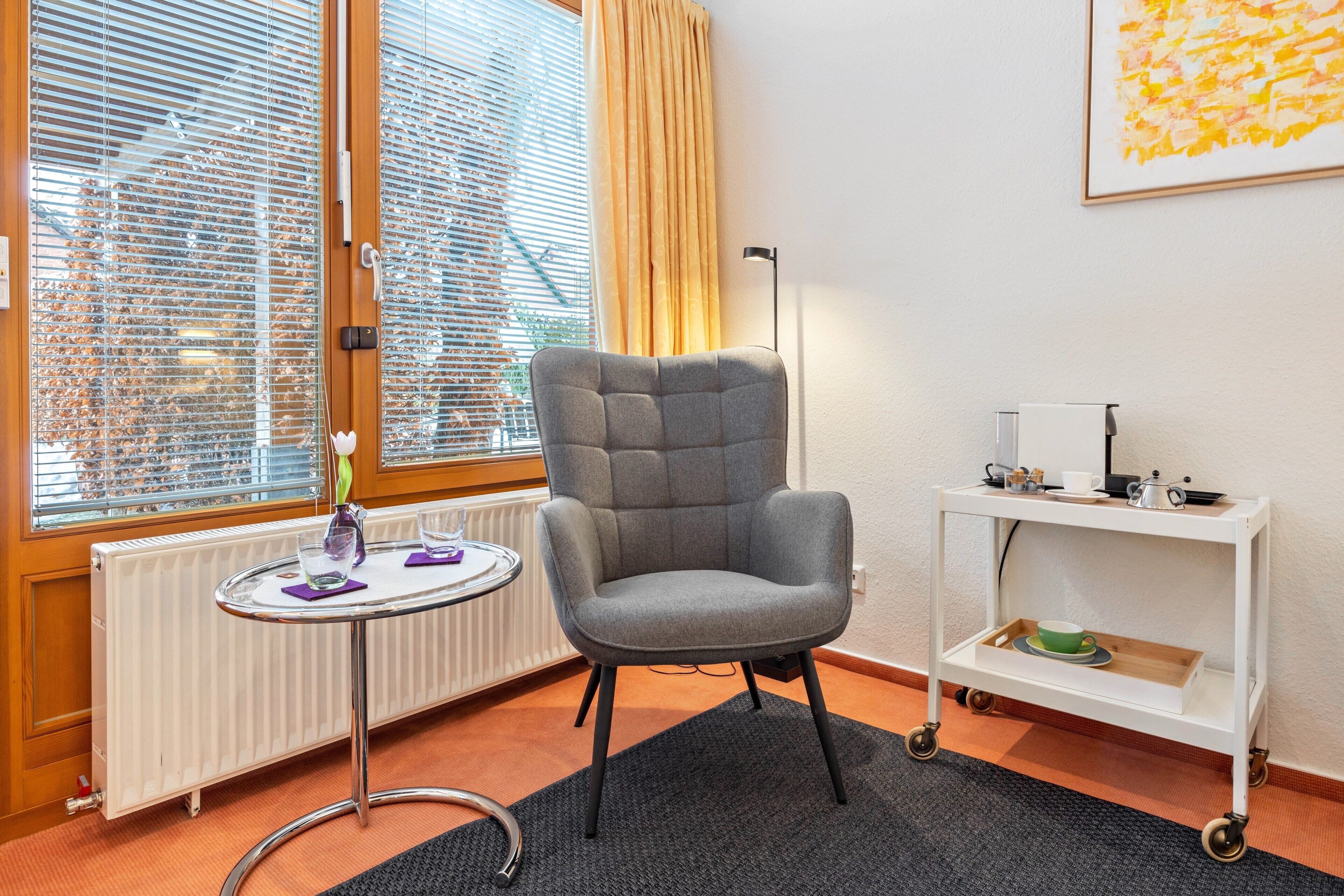 A room with a grey armchair, a small round table with a vase and glass, a white radiator, a window with blinds, a white shelf with a cup, and a framed picture on the wall.