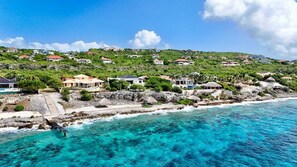 A coastal property with houses on a hillside, a pier extending into the sea, and clear turquoise waters. - Luxury seaside retreat with private beach (Sint Willibrordus)