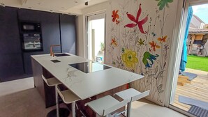 Una cocina moderna con encimera blanca y electrodomésticos integrados, una puerta corrediza que da a un balcón con terraza de madera y mobiliario exterior.