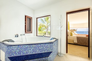 A bathroom with a large bathtub, a window with a view of palm trees, and a door leading to a balcony with a view of the ocean. - Elegant seaside retreat with private beach (Sint Willibrordus)