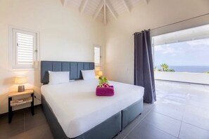 Un dormitorio con una cama grande, una mesita de noche, una ventana con persianas y vistas al océano.