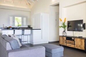 A modern living room with a grey sofa, a wooden cabinet with a TV, and a kitchen area with white cabinets and a refrigerator. - Modern sea-view villa with infinity pool (Sint Willibrordus)