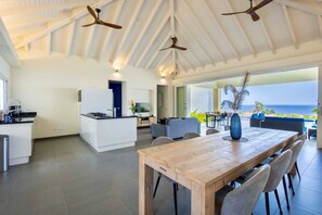 Una cocina amplia con una isla central, un comedor con mesa y sillas de madera, y un ventanal con vistas al océano.