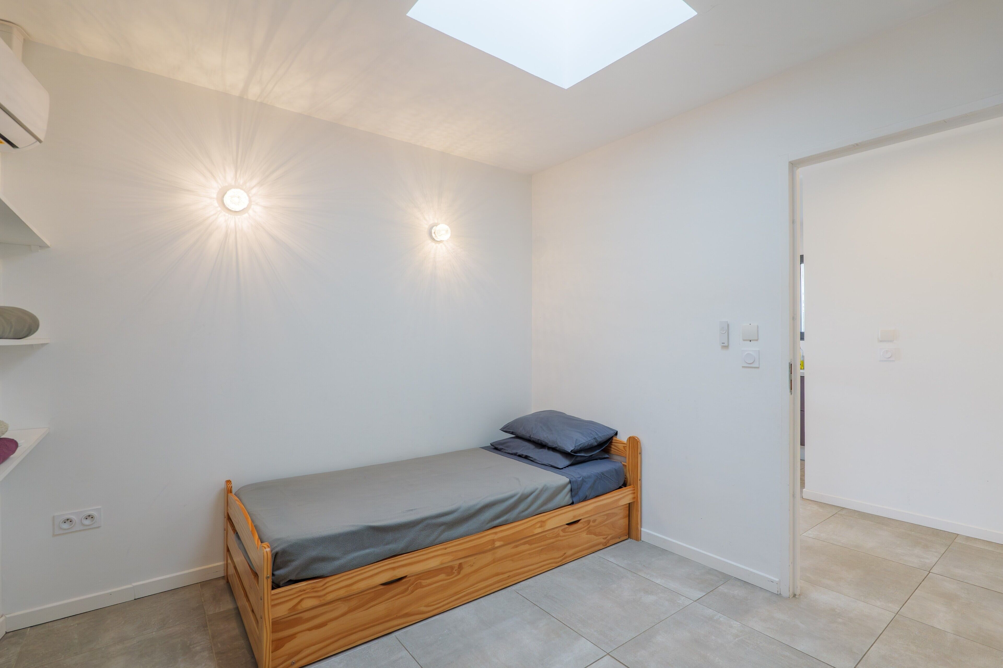Une petite chambre avec un lit simple, une lucarne, une porte blanche et une étagère avec quelques objets.