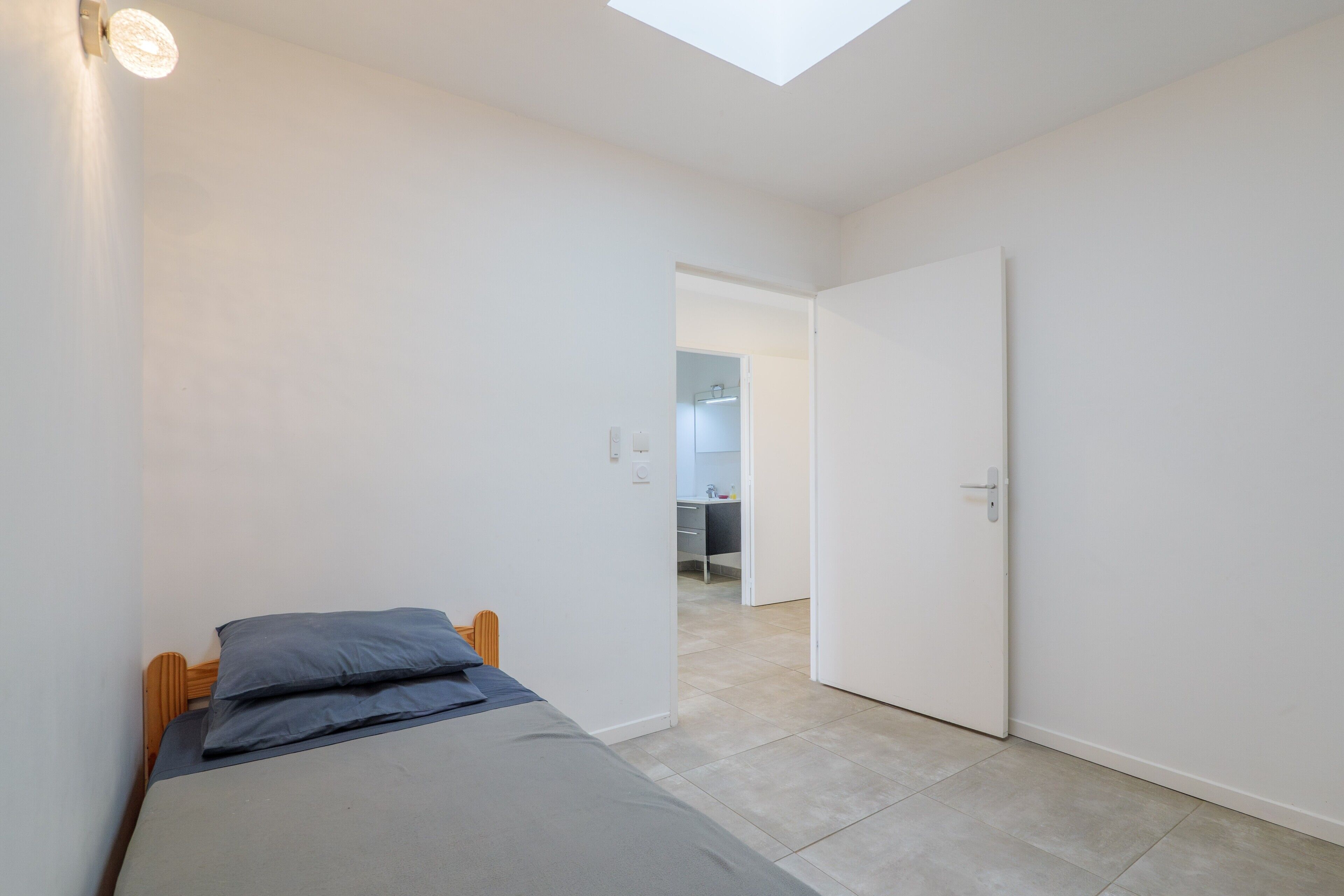 Une chambre à coucher comprenant un lit, une lucarne, une porte donnant sur une salle de bain et un couloir.