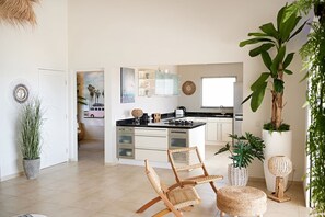 Une cuisine moderne avec des meubles blancs, un plan de travail noir et un îlot central. On aperçoit au premier plan des chaises en osier et une petite table.