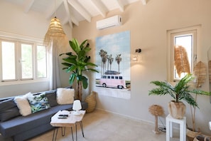 A living room with a grey sofa, a hanging light fixture, a large plant, a wall-mounted air conditioner, and a framed picture of a pink van. - Villa Caribbean Breeze 218 (Sint Willibrordus)