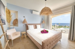 A bedroom with a large bed, a chair, a small stool, and a view of the ocean. - Villa Caribbean Breeze 218 (Sint Willibrordus)