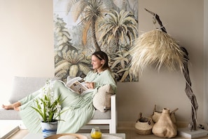 A person sitting on a sofa with a tropical-themed painting, a potted plant, and woven baskets on the floor. - Villa Caribbean Breeze 218 (Sint Willibrordus)