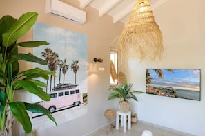 A room with a large window, a wall-mounted air conditioner, a hanging woven light fixture, a framed picture of a pink van, and a potted plant. - Villa Caribbean Breeze 218 (Sint Willibrordus)