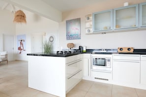A modern kitchen with white cabinets, a black countertop, and a central island. There is a painting of a flamingo on the wall, a hanging basket, and a coffee machine on the island. - Villa Caribbean Breeze 218 (Sint Willibrordus)