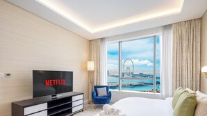 Ett hotellrum med en stor TV som visar "NETFLIX", en säng, en blå fåtölj, ett fönster med utsikt över ett pariserhjul och vatten, samt en lampa på väggen.